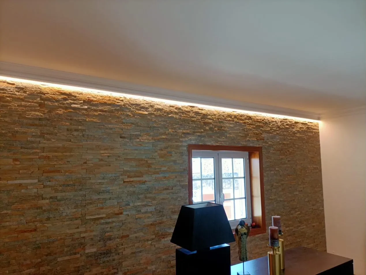 Parede revestida a pedra iluminada por fita LED na sanca, com candeeiro preto em primeiro plano